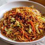 A close-up shot of savory, glossy fried noodles mixed with shredded cabbage and carrots, topped with dark sauce.