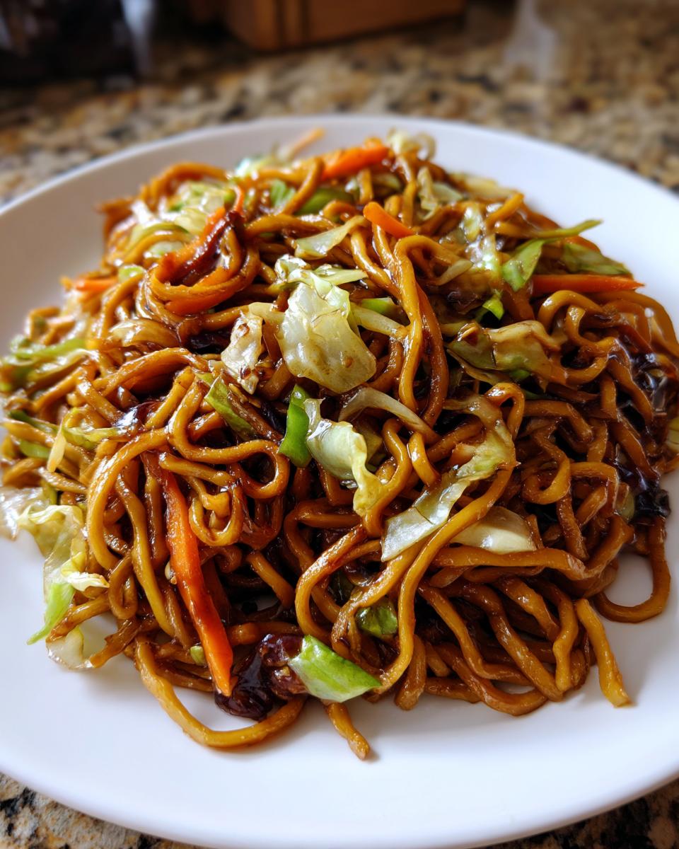 Close-up of a white plate piled high with glossy, savory fried noodles mixed with shredded cabbage and carrots.