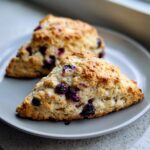 Two freshly baked, triangular blueberry scones with golden tops resting on a light grey plate.