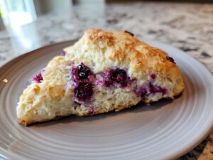 A close-up view of a freshly baked, triangular blueberry scone showing bright purple, juicy blueberries baked into the light pastry.