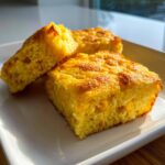 Three golden squares of moist cornbread casserole, studded with whole corn kernels, served on a white plate.