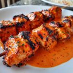 Close-up of grilled bang bang chicken skewers coated in a creamy, orange-red sauce on a white plate.