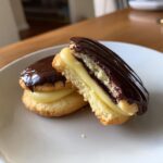 Two decadent boston cream pie cookies, one cut in half showing vanilla cream filling and chocolate glaze.