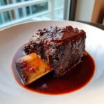 A single, rich, dark braised short ribs serving covered in thick sauce, resting in its juices.