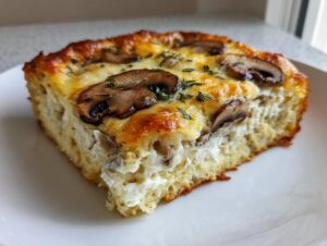 A square slice of baked breakfast casserole with cream of mushroom, topped with browned mushrooms and herbs.