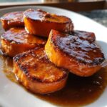 Five thick slices of candied sweet potatoes glistening with a rich, dark glaze on a white plate.