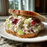 A thick, open-faced chicken salad sandwich featuring creamy chicken salad mixed with red grapes and green celery chunks on toasted bread.