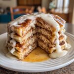 A tall stack of fluffy cinnamon roll pancakes, cut open to show cinnamon swirls, topped with thick white glaze.