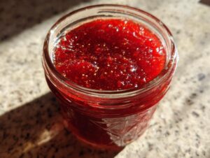 A close-up view of vibrant, glistening red christmas jam filling a glass canning jar, set on a speckled countertop.