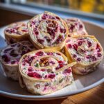 Five slices of cranberry tortilla roll ups filled with cream cheese, cranberries, and pecans, stacked on a white plate.
