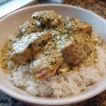 Close-up of creamy chicken and rice served in a white bowl, topped with herbs.