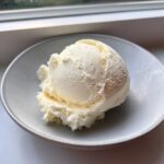 A single, perfectly scooped serving of creamy, pale yellow cottage cheese ice cream in a light grey bowl.