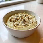 A close-up of a creamy, pale green dirty martini dip topped generously with crumbled blue cheese and a drizzle of liquid.