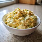 A close-up of creamy, yellow egg salad mixed with green celery and topped with fresh dill, served in a white bowl.