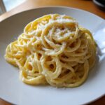 A close-up of perfectly twirled creamy lemon pasta topped with grated Parmesan cheese on a white plate.