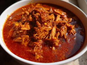 Close-up of a white bowl filled with vibrant red Pozole rojo, featuring tender shredded meat in a rich chili broth.