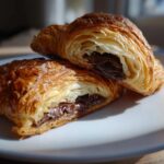 Close-up of a flaky, golden-brown chocolate croissant cut in half, revealing melted chocolate filling.