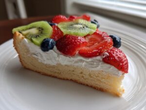 A close-up slice of fruit pizza sugar cookie topped with whipped cream, strawberries, kiwi, and blueberries.