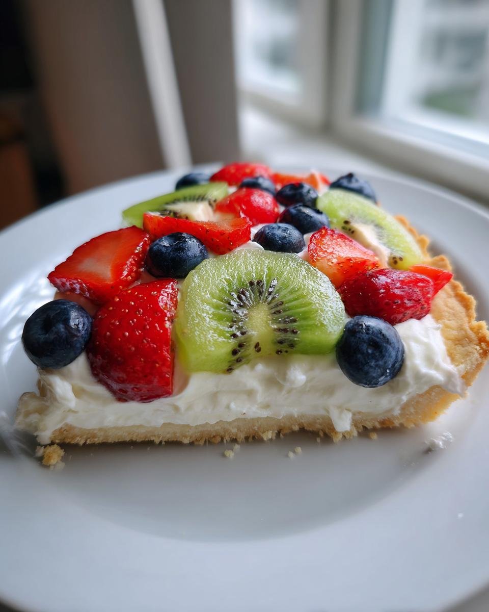 Close-up of a slice of fruit pizza sugar cookie topped with cream cheese frosting, strawberries, blueberries, and kiwi.