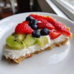 A close-up slice of fruit pizza sugar cookie topped with cream cheese frosting, strawberries, blueberries, and kiwi.