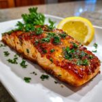 A perfectly cooked, glazed salmon fillet sprinkled with fresh parsley, served with a lemon wedge.