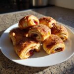 A small pile of freshly baked, golden brown Rugelach pastries dusted with cinnamon sugar on a white plate.