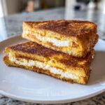 Two halves of golden brown stuffed french toast stacked on a white plate, showing a thick cream cheese filling.