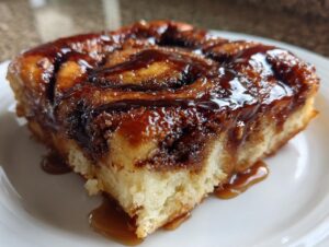 A close-up of a single, moist slice of cinnamon roll casserole topped with shiny caramel glaze.