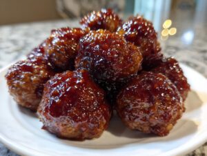 Close-up of several glistening Grape Jelly Meatballs piled high on a white serving plate.