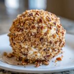 A perfectly formed Holiday cheese ball generously coated in chopped pecans, resting on a white plate.