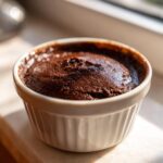 Close-up of rich, dark homemade chocolate pudding baked in a white fluted ramekin, catching sunlight.