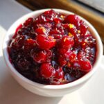 A close-up of chunky, homemade cranberry sauce in a white bowl, featuring whole cranberries and flecks of orange zest.