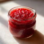Close-up of a small glass jar filled with glistening, deep red christmas jam, illuminated by bright sunlight.