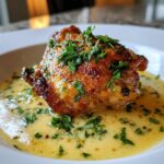 Close-up of a perfectly baked chicken thighs piece with crispy, golden-brown skin sitting in a pool of creamy yellow sauce and topped with fresh parsley.