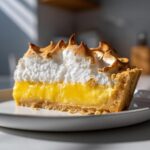 A close-up slice of lemon meringue pie showing bright yellow curd, crust, and peaks of toasted meringue.