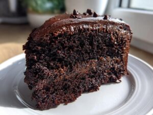 Close-up of a rich, dark slice of chocolate fudge cake with glossy frosting on a white plate.