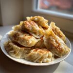 A close-up of a pile of golden-brown, pan-fried Traditional Chinese Dumplings stacked on a white plate near a window.