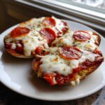 Two halves of a pepperoni english muffin pizza topped with melted mozzarella cheese and herbs on a white plate.