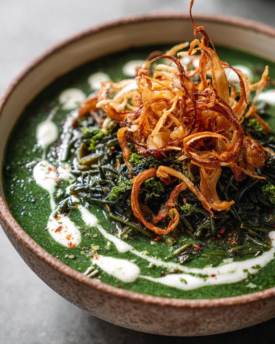 Close-up of a vibrant green persian noodle soup topped with fried onions and a swirl of white cream or yogurt.