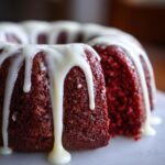 A close-up of a rich, dark red velvet bundt cake drizzled generously with thick white cream cheese glaze.