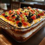 A layered Seven layer dip featuring visible layers of beans, sour cream, cheese, tomatoes, olives, and green peppers.