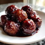 A close-up of several glistening Grape Jelly Meatballs coated in a thick, dark, sweet glaze on a white plate.