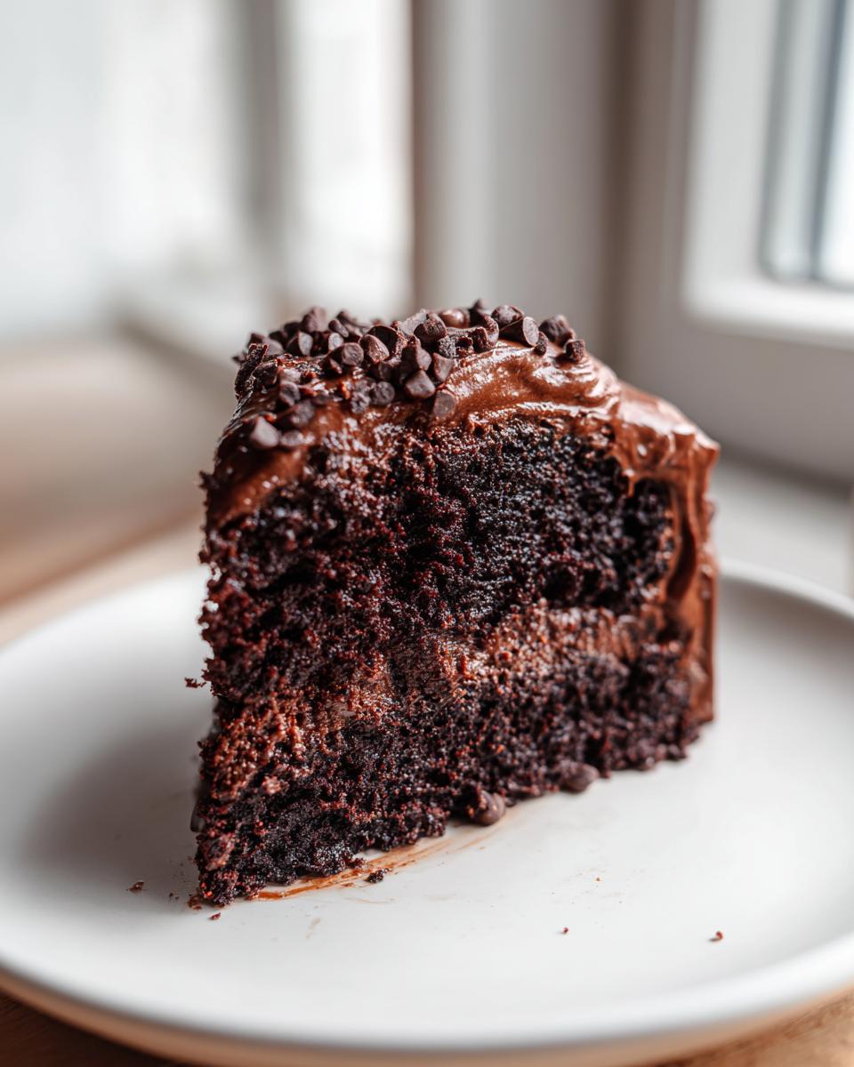 A close-up of a moist slice of chocolate fudge cake layered with frosting and topped with mini chocolate chips.