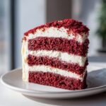 A tempting slice of multi-layered red velvet cake with thick white frosting layers, sitting on a white plate.