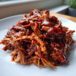 Close-up of shredded, saucy Slow Cooker Pulled Pork piled high on a white plate.