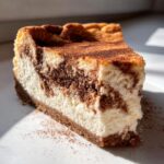 A close-up slice of snickerdoodle cheesecake showing a creamy white base swirled with cinnamon filling and topped with cinnamon dust.