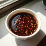 Close-up of a small white bowl filled with dark, rich Dumpling Sauce topped with visible chili flakes and garlic bits.
