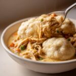 A spoonful lifts creamy shredded chicken and dumplings from a white bowl of rich chicken and dumplings stew.