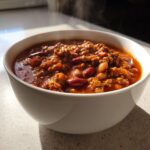 Close-up of a steaming white bowl filled with thick, hearty crockpot chili featuring ground meat and kidney beans.