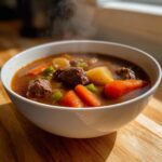A steaming white bowl filled with rich vegetable beef soup, featuring chunks of beef, carrots, and celery.